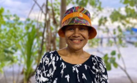 Woman in multicoloured hat smiling in front of trees looking to camera.