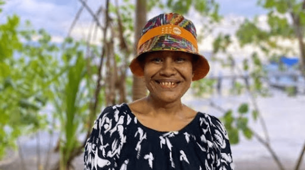 Woman in multicoloured hat smiling in front of trees looking to camera.