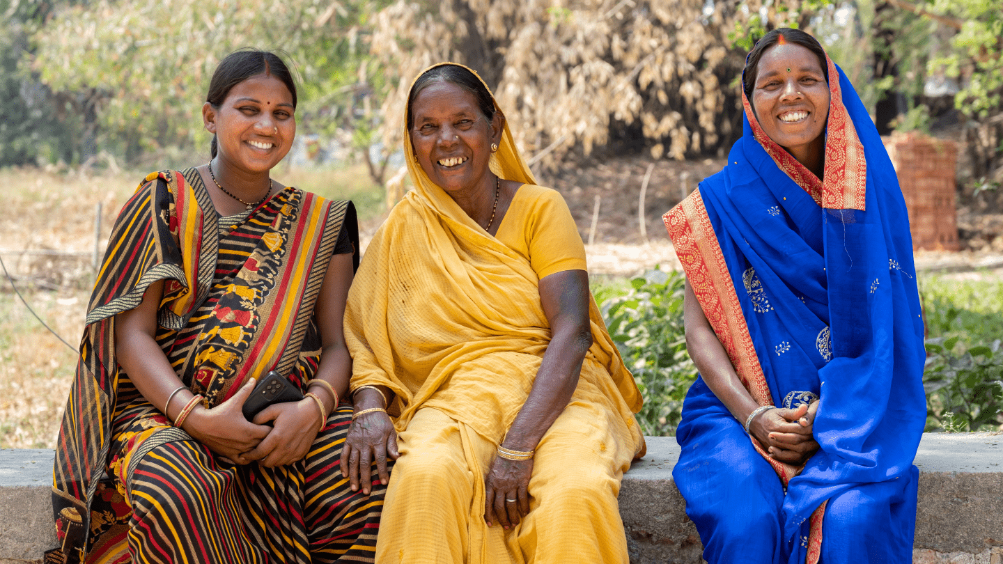 Three women from India smiling, they are sitting on a bench outdoors