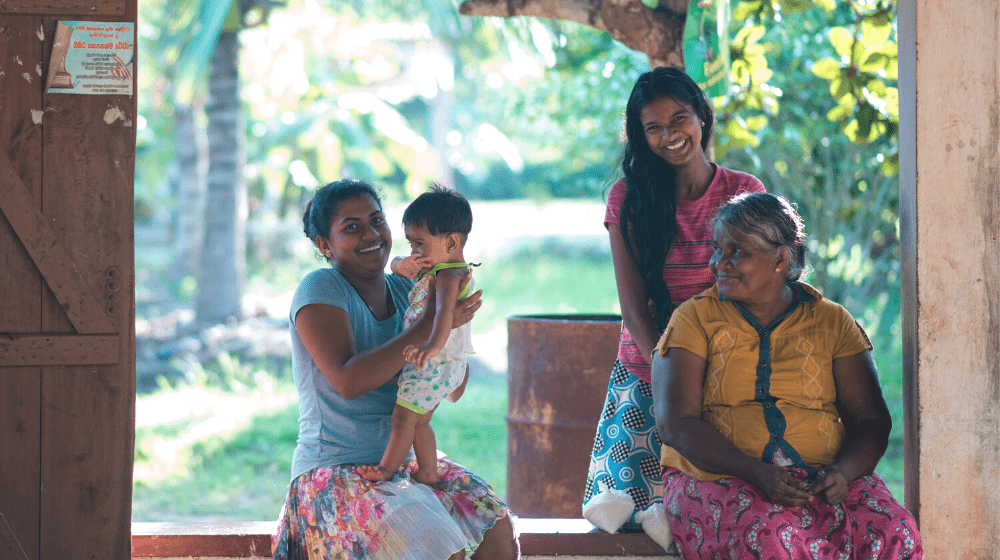 A lady with her newborn, her elder daughter, and her mother laugh while sitting outside their porch in Sri Lanka